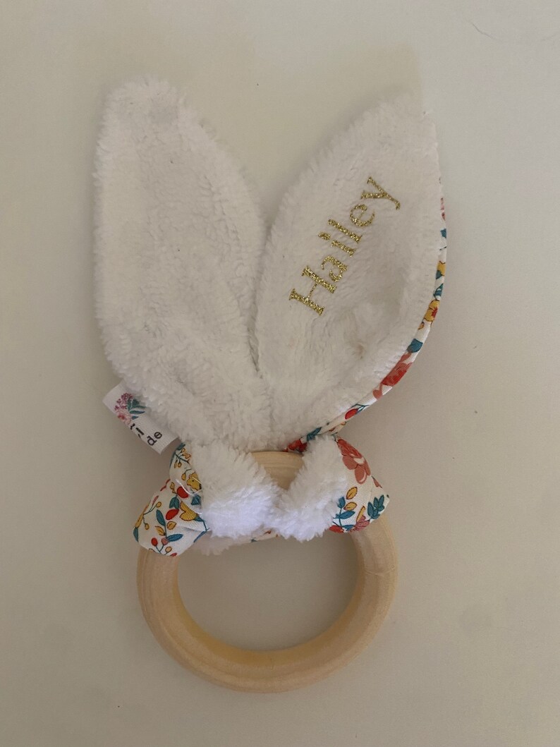 Pode incluir: Um mordedor de coelho branco com um anel de madeira e detalhes em tecido floral. As orelhas de coelho s&atilde;o macias e felpudas, e o anel &eacute; perfeito para as pequenas m&atilde;os agarrarem. O tecido &eacute; estampado com um padr&atilde;o floral colorido. As orelhas de coelho s&atilde;o bordadas com o nome "Hailey".