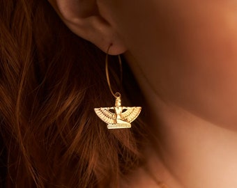 May include: Gold hoop earrings with a winged goddess charm. The charm is detailed and features a woman with outstretched wings.