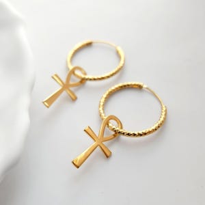 May include: Gold hoop earrings with a dangling ankh cross charm. The hoops are textured and the ankh is a simple design.