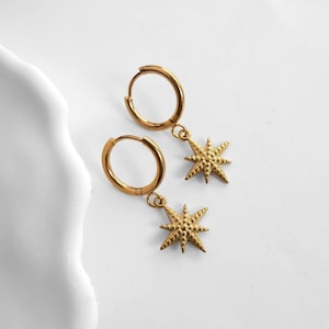 May include: A pair of gold-colored hoop earrings with star-shaped charms. The earrings are made of a shiny metal and feature a detailed star design. The earrings are displayed on a white surface.