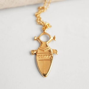 May include: A gold necklace with a pendant shaped like a stylized figure with geometric patterns. The pendant has a circular opening in the center.