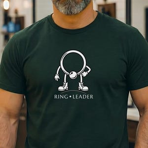 May include: Forest green t-shirt with a white graphic of a ring-shaped character with arms, legs, and sneakers. The text "RING LEADER" is below the graphic. The shirt is short-sleeved.