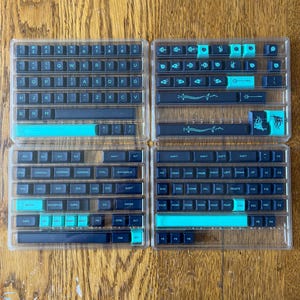 Puede incluir: Cuatro juegos de teclas de teclado en estuches de plástico transparente. Las teclas son negras y turquesas, con varios símbolos y letras. El diseño incluye teclas estándar, teclas de función y teclas especiales como "Enter" y "Shift".