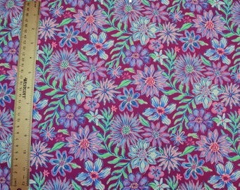 Op de afbeelding: Een close-up van een stof met een bloemenpatroon in tinten blauw, paars en roze op een bordeauxrode achtergrond. De stof is bedekt met verschillende bloemen, waaronder madeliefjes en kleinere bloemen.