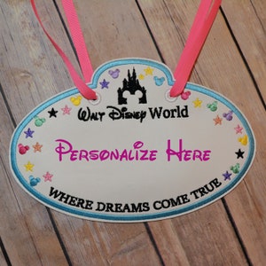 May include: White oval name tag with blue trim, featuring a black silhouette of the Disney castle, stars, and Mickey Mouse heads. The text "Walt Disney World" and "Where Dreams Come True" are embroidered in black. The text "Personalize Here" is in pink.