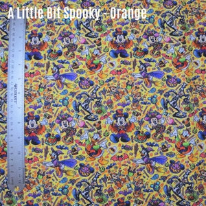 May include: A yellow fabric with a Halloween-themed print featuring Mickey Mouse, Donald Duck, Goofy, and other Disney characters. The print also includes candy, bats, pumpkins, and other Halloween imagery. The text "A Little Bit Spooky - Orange" is printed at the top of the fabric.