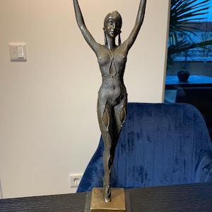 May include: Bronze sculpture of a woman with arms raised. The figure has a detailed, textured surface and is mounted on a square base. The sculpture is approximately 46 cm tall.