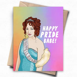 May include: Greeting card featuring a cartoon illustration of a woman in a blue dress and tiara, with the text "HAPPY PRIDE BABE!". The card has a rainbow gradient background and is placed on a brown envelope.