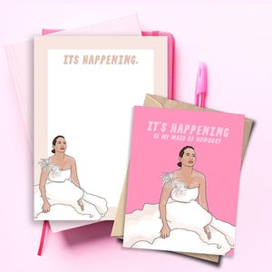 May include: A pink greeting card and a notepad featuring an illustration of a person in a white strapless dress with feather detail. The card text reads IT'S HAPPENING BE MY MAID OF HONOUR? and the notepad text reads IT'S HAPPENING. A pink pen is included.