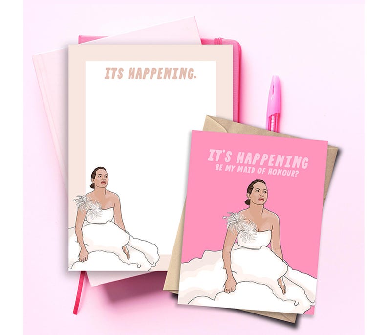 May include: A pink greeting card and a notepad featuring an illustration of a person in a white strapless dress with feather detail. The card text reads IT'S HAPPENING BE MY MAID OF HONOUR? and the notepad text reads IT'S HAPPENING. A pink pen is included.