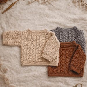 Puede incluir: Tres suéteres de punto trenzado en beige, gris y marrón se exhiben sobre una tela de tono neutro. Los suéteres tienen un cuello redondo clásico y mangas texturizadas. También se ven hilo, un ganchillo y unas tijeras.