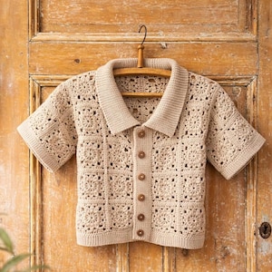 Puede incluir: Camisa de crochet beige de manga corta con cuello y botones de madera. La camisa está hecha con un patrón cuadrado y cuelga de una percha de madera contra una puerta de madera.