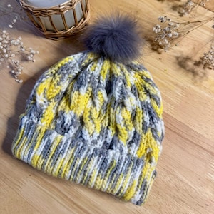 May include: A handmade knit beanie in yellow, gray, and white with a gray faux fur pom-pom. The hat features a cable knit design and is displayed on a wooden surface. A candle in a wicker-wrapped jar and a ball of yarn are also visible.