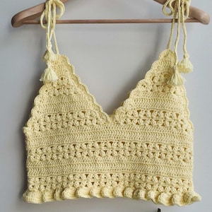May include: A light yellow, handmade crochet top with a V-neck design. The top features a scalloped edge and decorative tassels on the straps. The garment is displayed on a wooden hanger against a white background.