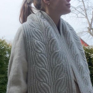 May include: A light gray knitted scarf with a leaf pattern. The scarf is wrapped around the neck and shoulders. The knitted pattern is visible, with a ribbed texture. The scarf is made of a soft, warm material.