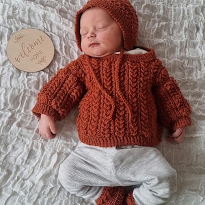 May include: A newborn baby wearing a rust-colored knitted hooded sweater, matching booties, and a hat. The baby is also wearing gray ribbed pants. A wooden disc with the words "welcome home" is next to the baby.