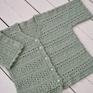 May include: A sage green, crocheted cardigan with a V-neck and long sleeves. The cardigan features a scalloped edge and is fastened with four pearl-like buttons. The openwork design adds a delicate touch to the handmade garment.