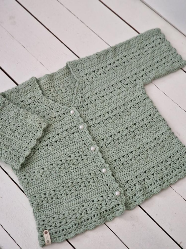 May include: A sage green, crocheted cardigan with a V-neck and long sleeves. The cardigan features a scalloped edge and is fastened with four pearl-like buttons. The openwork design adds a delicate touch to the handmade garment.