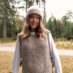 May include: A woman wearing a brown cable knit vest with a white long-sleeved shirt underneath. She is also wearing a brown beanie and brown trousers. The photo was taken outdoors in a woodland setting.