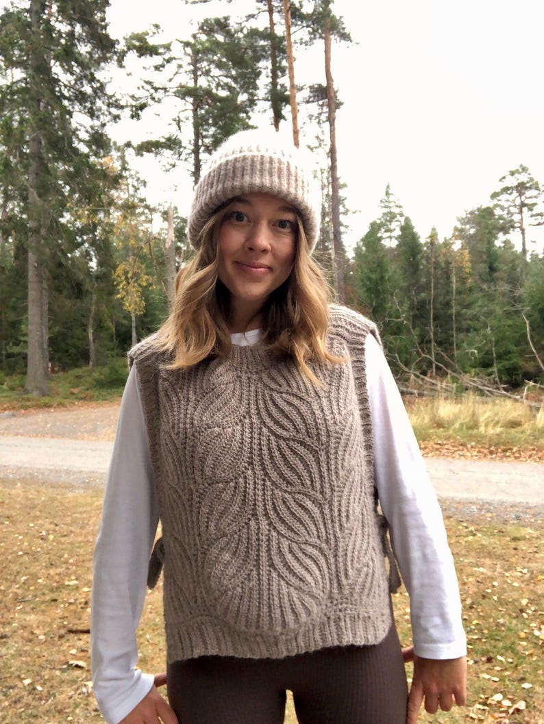 May include: A woman wearing a brown cable knit vest with a white long-sleeved shirt underneath. She is also wearing a brown beanie and brown trousers. The photo was taken outdoors in a woodland setting.