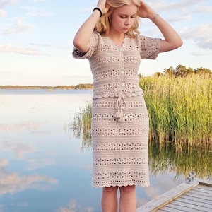 May include: A beige crochet dress with short sleeves, a button-up front, and a drawstring waist. The dress is knee-length and has a scalloped hem. The person is standing on a wooden dock near a body of water.
