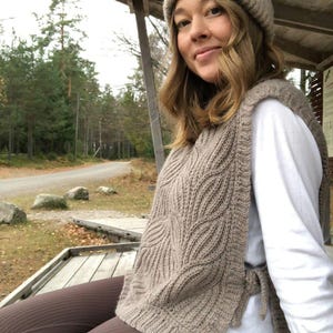 Könnte beinhalten: Eine Frau trägt eine braune Strickweste mit einem Krawattendetail, ein weißes Hemd und eine braune Hose. Sie trägt eine braune Strickmütze und sitzt auf einem Holzdeck.