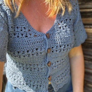 May include: A light blue crochet cardigan with short sleeves and a V-neck. The cardigan has wooden buttons and a scalloped hem. The crochet pattern features openwork details. The garment is paired with blue jeans.