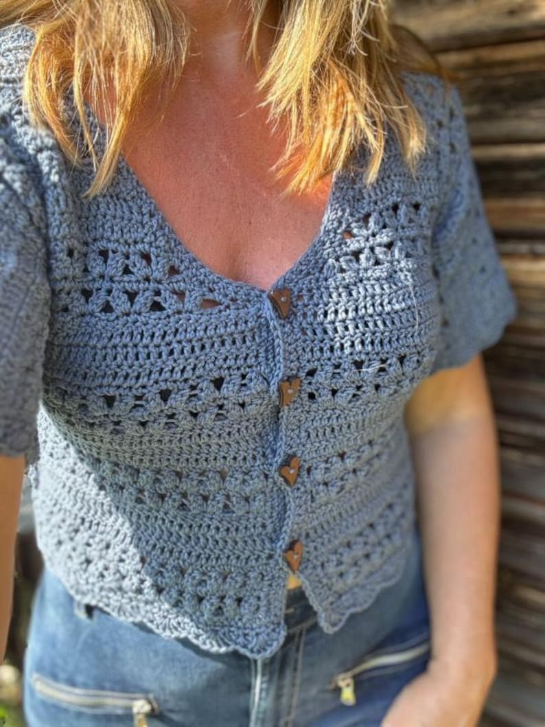 May include: A light blue crochet cardigan with short sleeves and a V-neck. The cardigan has wooden buttons and a scalloped hem. The crochet pattern features openwork details. The garment is paired with blue jeans.