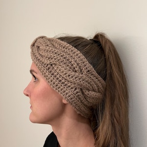 Peut inclure: Un bandeau en crochet marron avec un motif en tricot torsadé. Le bandeau est porté par une personne aux cheveux longs et bruns.