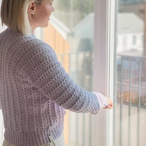 以下が含まれることがあります： 白の縁取りのある薄い紫色のクロシェ編みのカーディガンを着た女性。カーディガンは短く、ゆったりとしたフィット感です。女性は窓の近くに立って外を見ています。