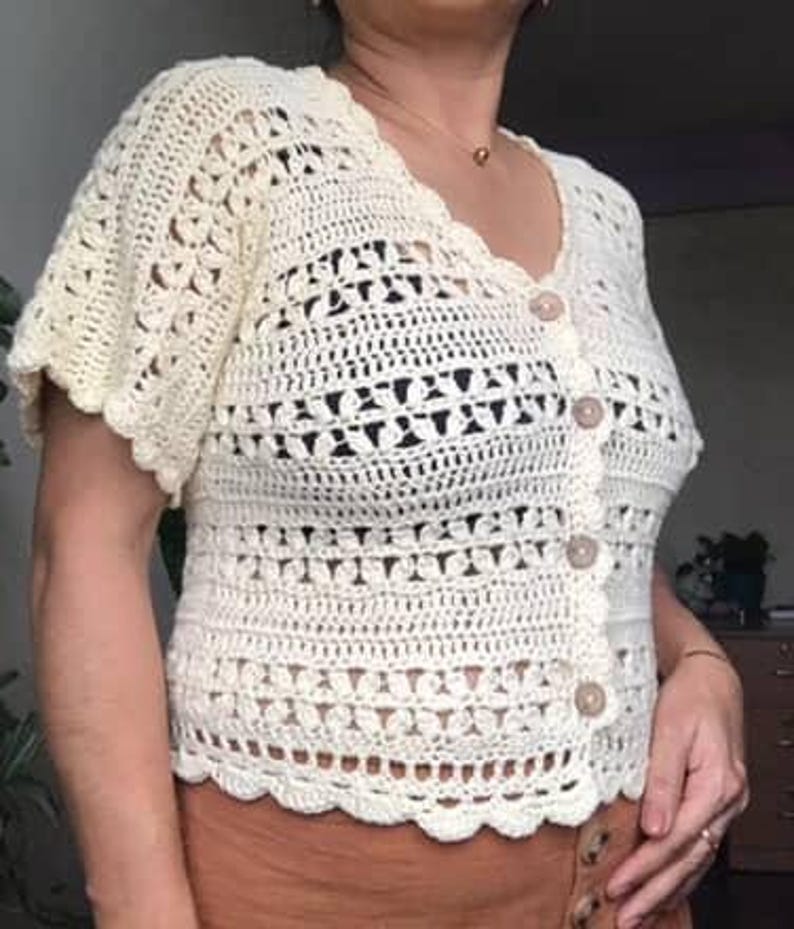 May include: Cream-colored, short-sleeved crochet cardigan with a scalloped edge and button closure. The handmade top features a detailed openwork design and is made of a soft, textured yarn. A stylish and unique addition to any wardrobe.