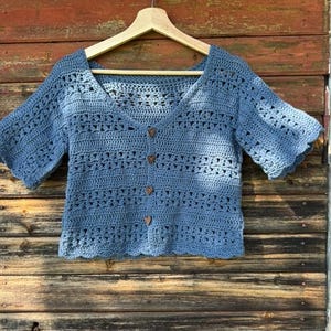May include: A blue crochet top with short sleeves and a V-neck. The top has a scalloped hem and is buttoned with wooden heart-shaped buttons. The garment is displayed on a wooden hanger against a weathered wooden background.