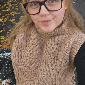 May include: A close-up of a person wearing a brown knitted jumper with a leaf pattern. The jumper has a high neck and is made of a soft, textured yarn.