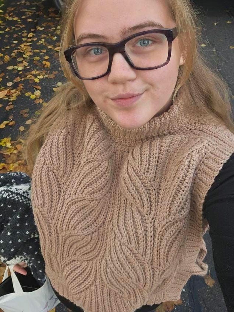 May include: A close-up of a person wearing a brown knitted jumper with a leaf pattern. The jumper has a high neck and is made of a soft, textured yarn.