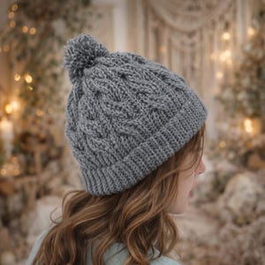 以下が含まれることがあります： グレーのケーブル編みのビーニー帽。上部にポンポンが付いています。帽子は、織り模様と折り返しのつばが特徴です。ビーニーは柔らかく暖かい素材でできており、寒い季節に最適です。長いウェーブのかかった茶色の髪の人がかぶっています。