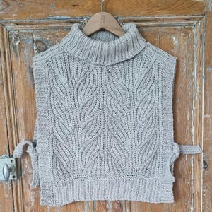 May include: A grey knitted turtleneck sweater vest with a leaf pattern. The vest has ties on the sides.