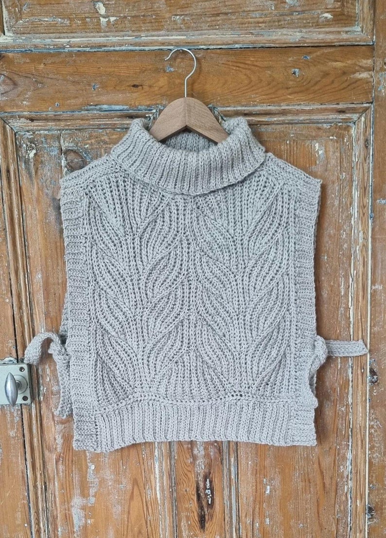 May include: A grey knitted turtleneck sweater vest with a leaf pattern. The vest has ties on the sides.