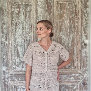 May include: A beige crochet two-piece set, consisting of a short-sleeved button-up top and a knee-length skirt. The outfit features a delicate openwork pattern and a drawstring detail at the waist. The model is standing in front of a weathered wooden door.