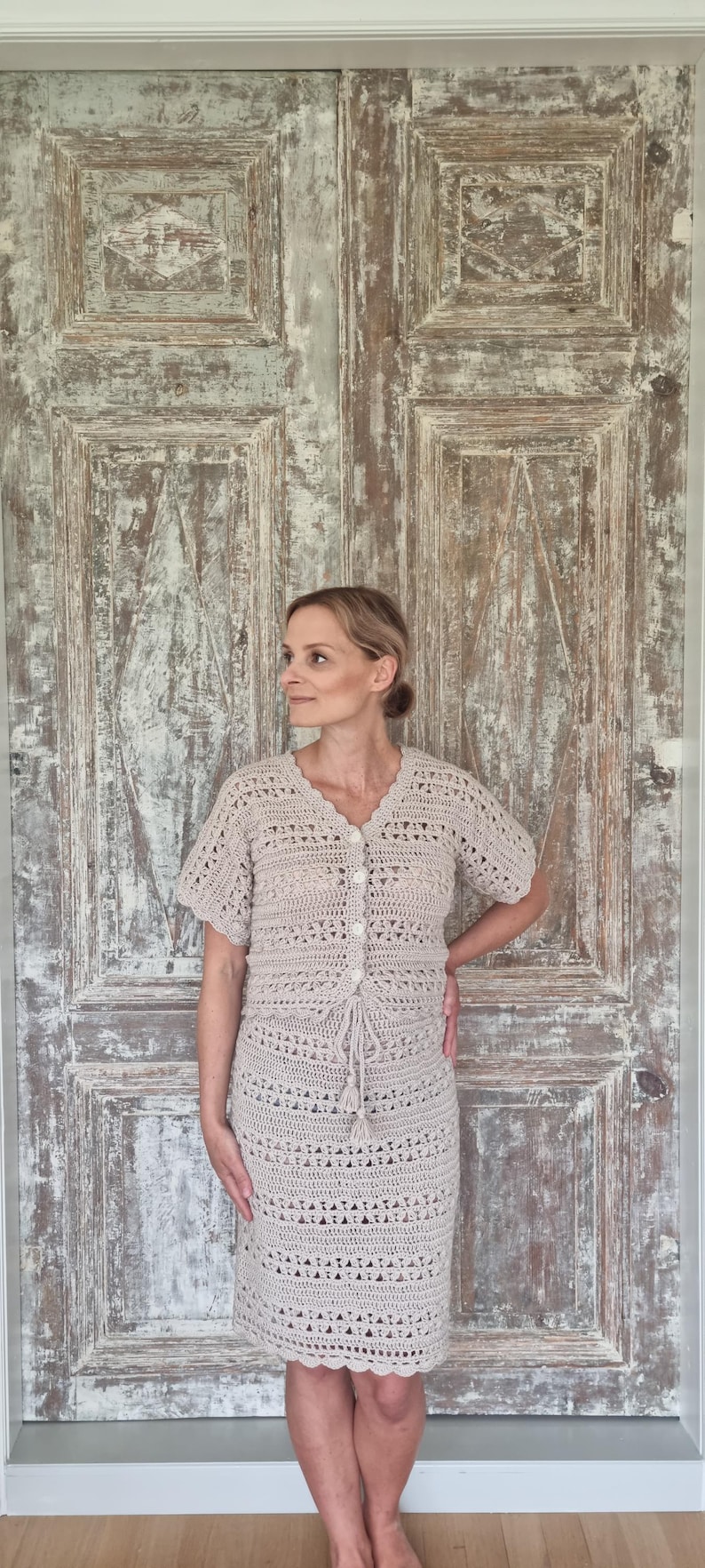 May include: A beige crochet two-piece set, consisting of a short-sleeved button-up top and a knee-length skirt. The outfit features a delicate openwork pattern and a drawstring detail at the waist. The model is standing in front of a weathered wooden door.