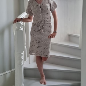 May include: A beige, two-piece crochet outfit, consisting of a short-sleeved top with button closures and a knee-length skirt. The outfit features a drawstring at the waist and is worn on a staircase.