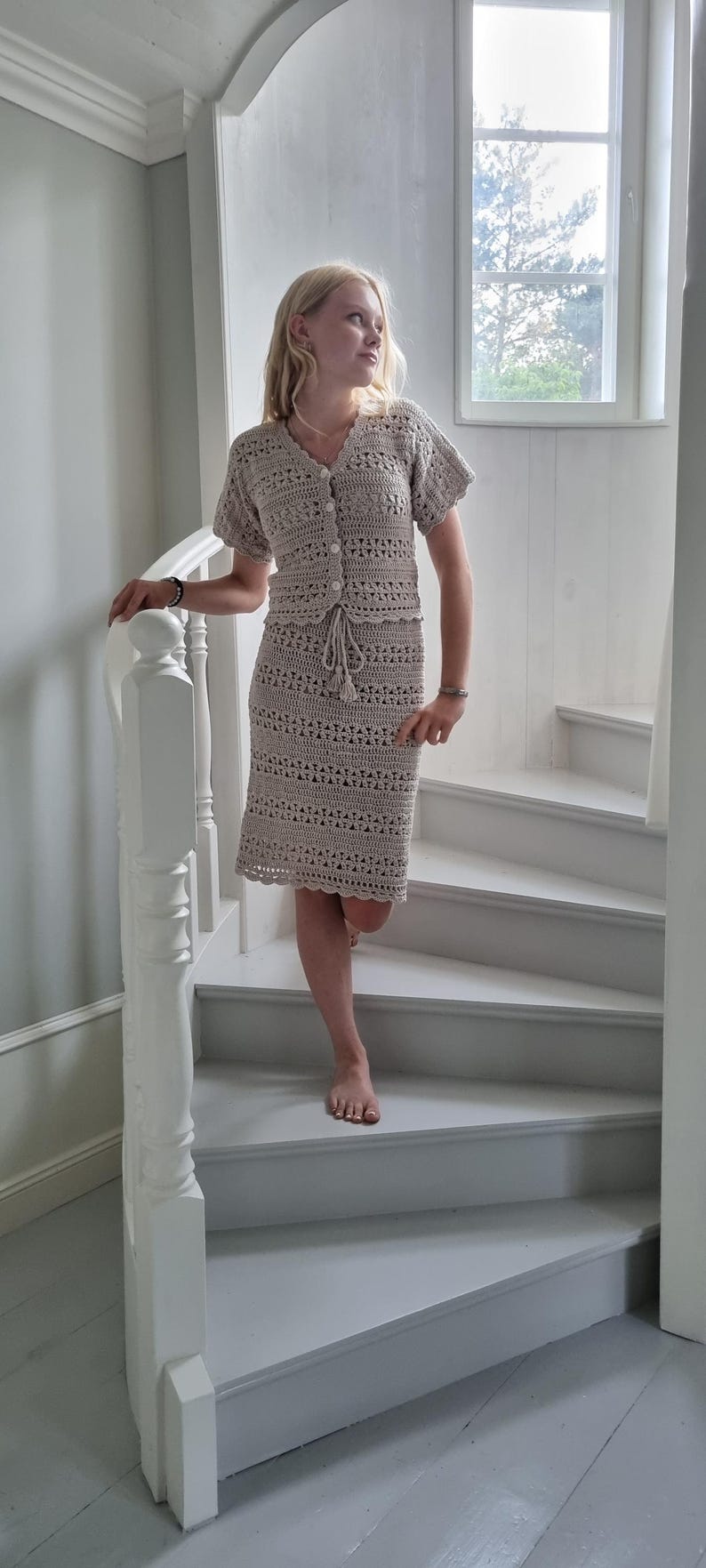 May include: A beige, two-piece crochet outfit, consisting of a short-sleeved top with button closures and a knee-length skirt. The outfit features a drawstring at the waist and is worn on a staircase.