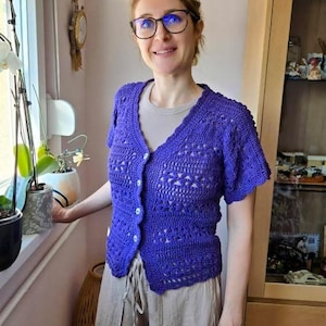 May include: A woman wearing a short-sleeved, button-up, purple crochet cardigan. The cardigan has a V-neck and a textured pattern. She is also wearing a light-colored shirt and tan pants. The background includes a window, plants, and a wooden cabinet.