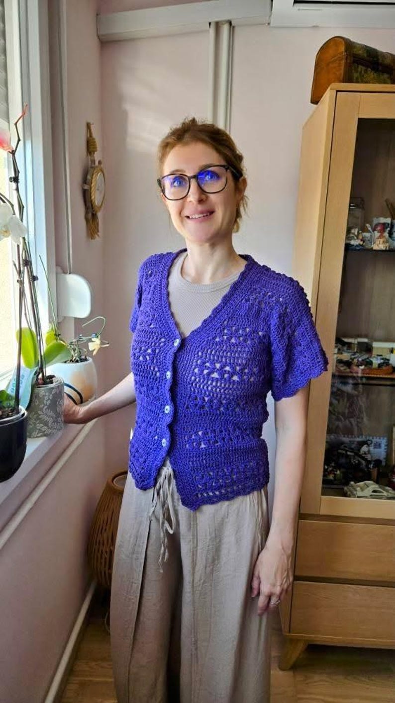 May include: A woman wearing a short-sleeved, button-up, purple crochet cardigan. The cardigan has a V-neck and a textured pattern. She is also wearing a light-colored shirt and tan pants. The background includes a window, plants, and a wooden cabinet.