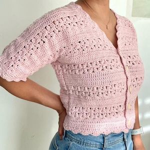 May include: A light pink, short-sleeved crochet cardigan with a V-neck and button closure. The crochet pattern features openwork and textured stitches. The edges of the cardigan have a scalloped design.