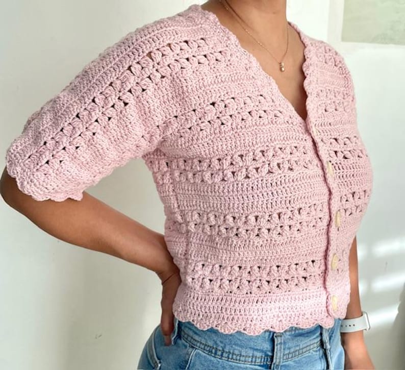 May include: A light pink, short-sleeved crochet cardigan with a V-neck and button closure. The crochet pattern features openwork and textured stitches. The edges of the cardigan have a scalloped design.
