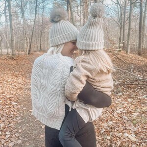 May include: A woman and a child wearing matching grey knit hats with pom-poms. The woman is wearing a white cable knit sweater and black leggings. The child is wearing a beige fleece jacket and black leggings.