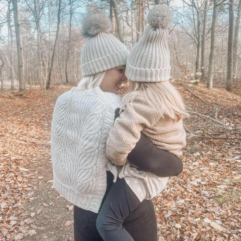 May include: A woman and a child wearing matching grey knit hats with pom-poms. The woman is wearing a white cable knit sweater and black leggings. The child is wearing a beige fleece jacket and black leggings.