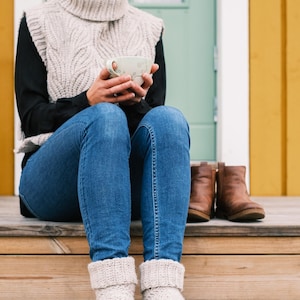May include: A woman wearing a beige knitted sweater vest, blue jeans, and white knitted socks sits on a wooden porch with a cup of coffee in her hands.