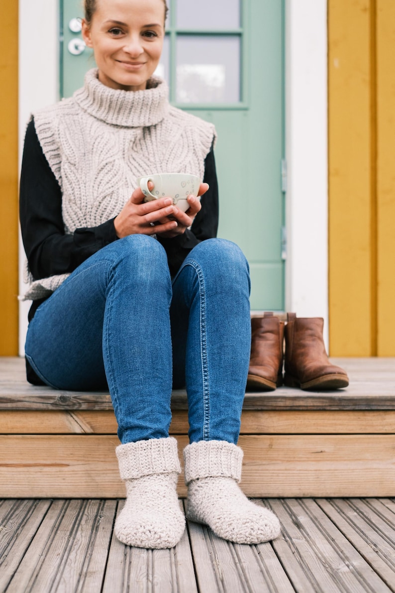 May include: A woman wearing a beige knitted sweater vest, blue jeans, and white knitted socks sits on a wooden porch with a cup of coffee in her hands.