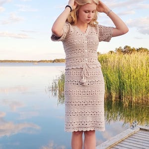 May include: A beige crochet dress with a matching short-sleeved top and knee-length skirt. The dress features a button-down front and a drawstring waist. The person is standing barefoot on a wooden dock near a body of water.